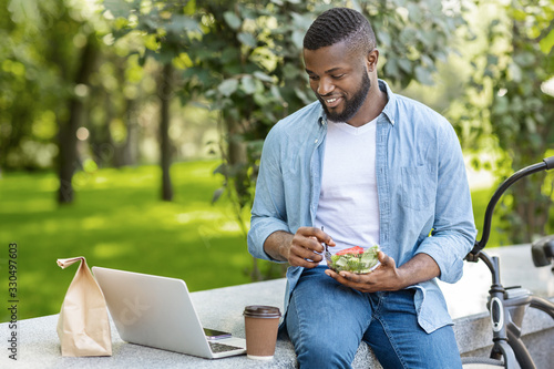 Black Guy Eating Salad And Drinking Coffee Outdoors, Relaxing With Laptop