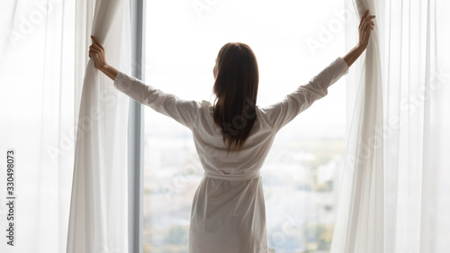 Back rear view confident woman in night robe gown pulling opening lace curtains after waking up in morning, admiring panorama view near big window, enjoying start of new day at home or hotel.