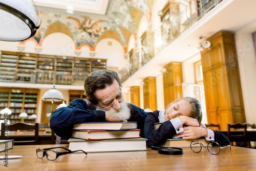 Tired senior bearded man teacher professor and his student or granddaughter are sleeping in a library liying on the table with a lot of books in old vintage library photo