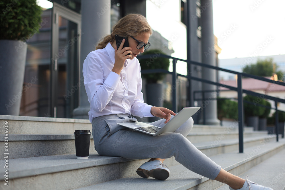 Smiling blonde woman using laptop and talking by smartphone while sitting outdoors. Smiling blonde woman using laptop and talking by smartphone while sitting outdoors.
