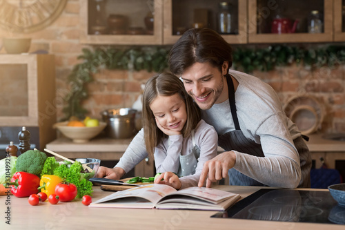 Fotografi Happy dad and little daughter checking recipe in cookbook together