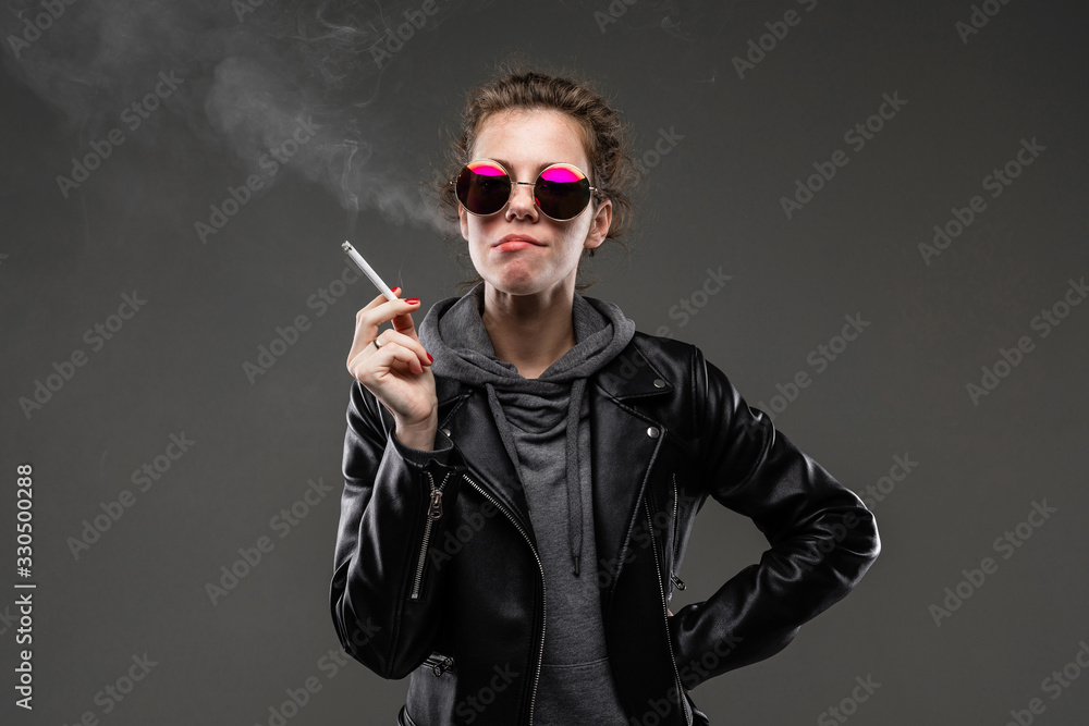 A young girl with rough facial features in a black jacket smokes isolated on black background