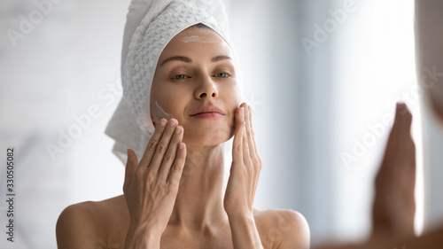 Closeup head shot pleasant beautiful woman applying moisturizing creme on face after shower. Smiling young pretty lady wrapped in towel smoothing perfecting skin, daily morning routine concept.