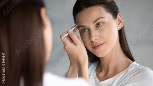 Tableau sur toile Mirror reflection attractive young brunette woman using tweezers, doing correction procedure in bathroom, satisfied with long lasting styling or perfect eyebrows shape, face treatment concept