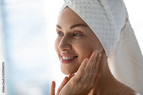 Close up head shot beautiful happy young woman with towel on head looking away in mirror, touching facial skin. Smiling attractive lady doing morning skincare routine in bathroom after showering.