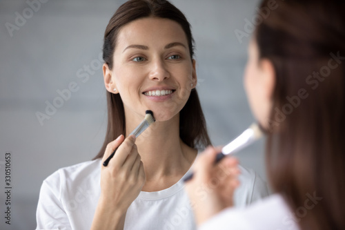 Head shot close up attractive smiling 30s woman using soft brush  applying foundation on face. Happy brunette lady enjoying doing daily morning make up routine  preparing for workday near mirror.