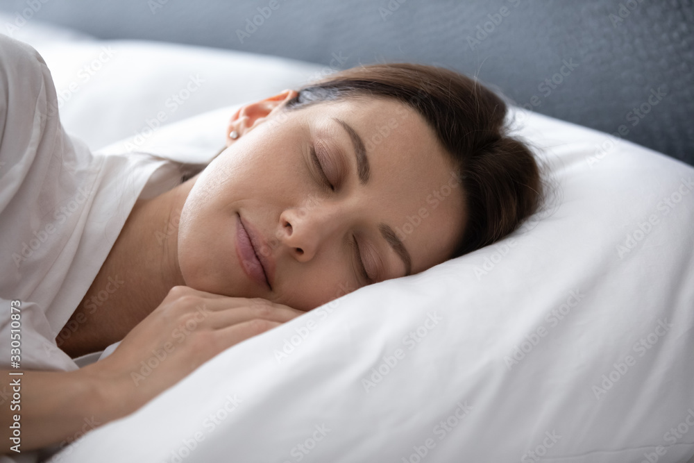 Close up head shot tranquil peaceful young woman enjoying good night rest on soft pillow in bedroom at home. Calm pleasant brunette lady seeing sweet dreams, sleeping alone in cozy bed in hotel.