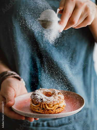 Woman sprinkles icing sugar on Choux Paris Brest pastry with hazelnut