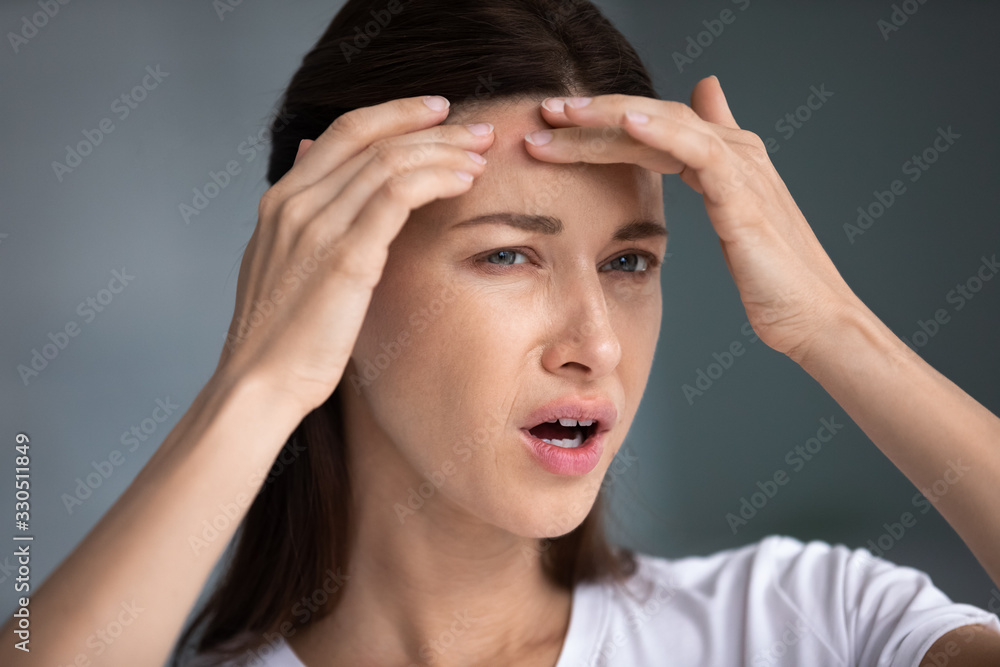 Close up head shot stressed frustrated young brunette woman looking in mirror, touching forehead, worrying about skin problems, wrinkles acne pimples, annoyed by blackheads, cosmetology treatment. Close up head shot stressed frustrated young brunette woman looking in mirror, touching forehead, worrying about skin problems, wrinkles acne pimples, annoyed by blackheads, cosmetology treatment.