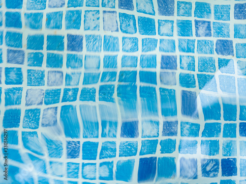 The light reflected on the top of the water surface of the pool lined with blue mosaic tiles below. Water ripples on blue-tiled swimming pool background.