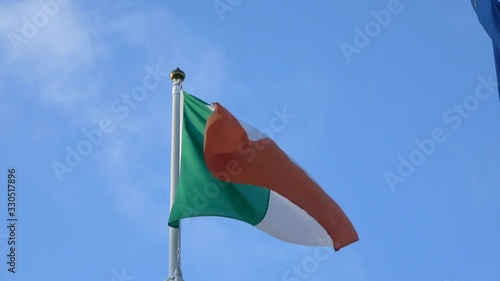 4K. Ireland's flag waving in the wind on flagpole at Dublin city. Banner flutters at day in front of blue sky. Close up of a real fabric texture ensign fluttering.