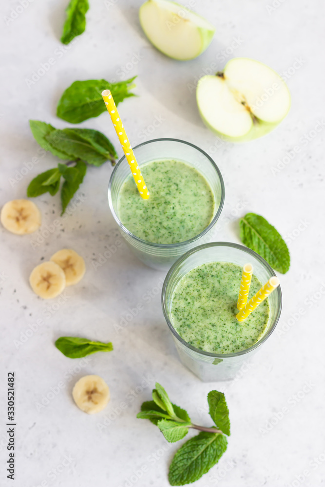 Two glasses with green spinach, banana, apple and mint smoothie with yellow straw. Light grey stone background. Healthy food concept. Two glasses with green spinach, banana, apple and mint smoothie with yellow straw. Light grey stone background. Healthy food concept.