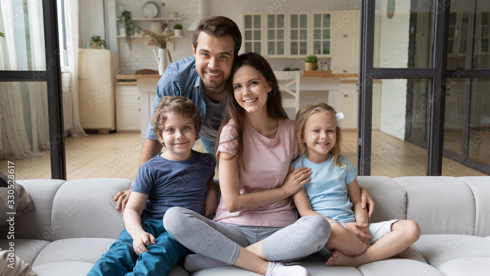 Portrait of happy successful young family with children first time home buyers sit on comfortable couch, smiling parents and little kids relax in living room look at camera posing, ownership concept Portrait of happy successful young family with children first time home buyers sit on comfortable couch, smiling parents and little kids relax in living room look at camera posing, ownership concept