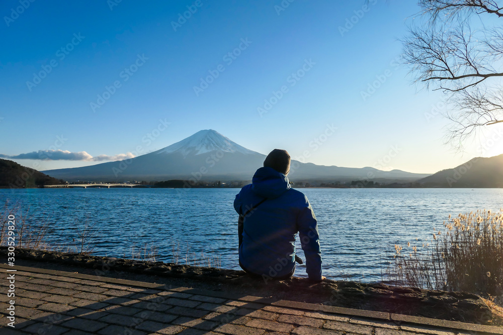 A man sitting at the side of Kawaguchiko Lake and watching Mt Fuji, Japan. The mountain surrounded by clouds, top of it covered with snow. Exploring new places. Calmness. Soft sunset colors