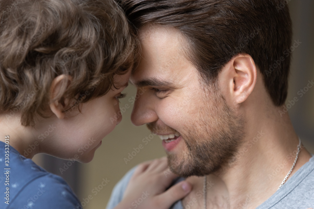 Fototapeta premium Close up of smiling young dad and cute small son touch foreheads look in eyes enjoying sweet tender moment together, happy father and little preschooler boy child embrace cuddle showing love