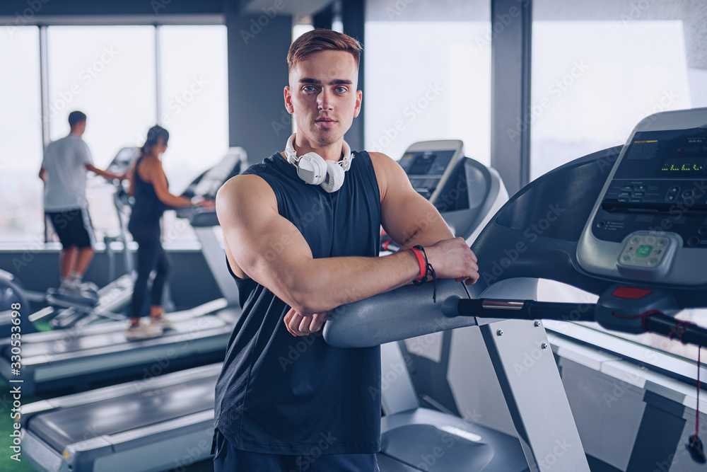 Smiling positive confident male personal instructor with arms crossed arms near treadmill at gym in fitness gym. Smiling positive confident male personal instructor with arms crossed arms near treadmill at gym in fitness gym.