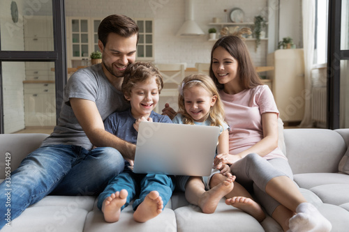 Overjoyed young family with small children sit relax on comfortable couch at home enjoying funny video on laptop, happy parents rest with kids on cozy sofa watching movie using computer together
