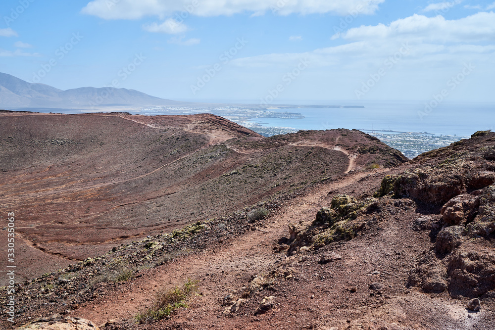 Der Vulkan Montana Roja de Playa Blanca mit einer Höhe von 194m auf der Südwestspitze der Kanareninsel Lanzarote und im Hintergrund die Feuerberge
