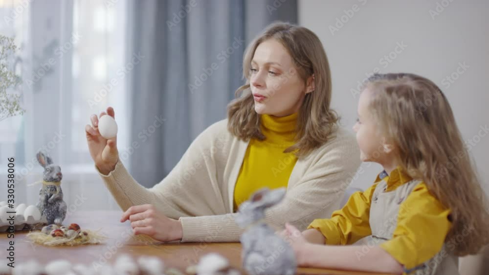 Handheld shot of beautiful young mother holding white boiled egg and talking to cute little girl. Easter bunny figurines standing on table Handheld shot of beautiful young mother holding white boiled egg and talking to cute little girl. Easter bunny figurines standing on table
