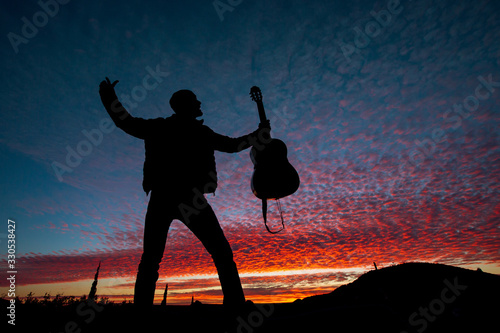 Silhouette of male musician standing, holding guitar on a scenic violet sunset sky 