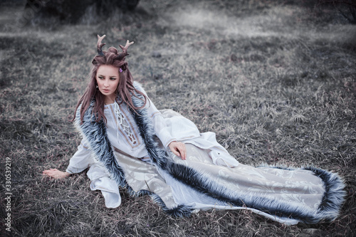Portrait of amazing cute young woman in fairy tale image with long hair in interesting hairstyle and horns on her head in beige dress with gray fur sitts on grass