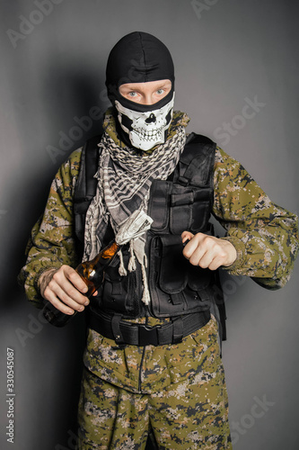 A man in a balaclava with a skull pattern, dressed in camouflage with a hood and a bulletproof vest, is holding a Molotov cocktail in his hands. A protester throws a Molotov cocktail. Studio photo on 
