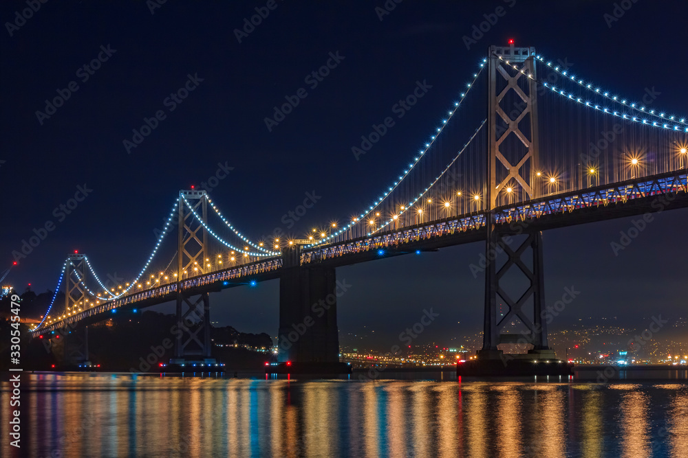 San Francisco Bay Bridge at night, lit up by yellow and blue lights, reflecting of the water in the Bay, long exposure
