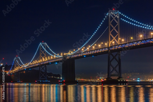San Francisco Bay Bridge at night  lit up by yellow and blue lights  reflecting of the water in the Bay  long exposure