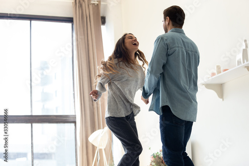Happy loving couple having fun, laughing and jumping, excited young woman and man dancing at home together, celebrating success, relocation or anniversary, funny family activity in modern room