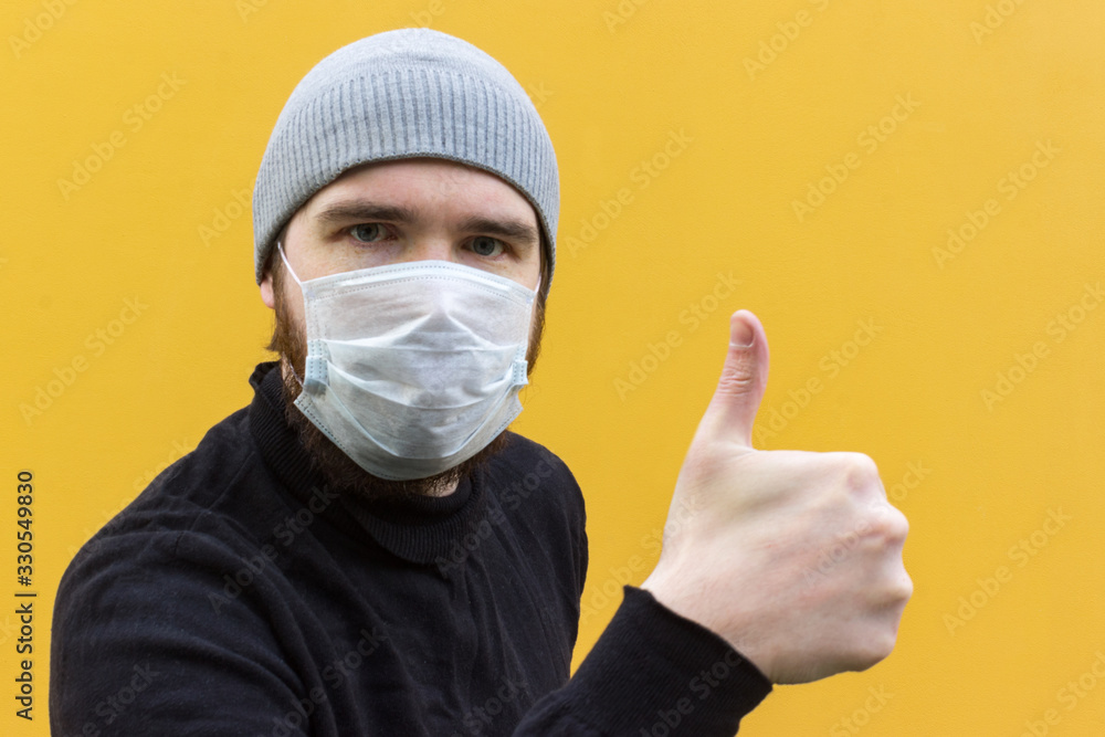 a man in a black jacket wearing a disposable mask to protect against coronavirus. Yellow background, emotions on the face. Person in a grey hat. Safety measures in case of illness. a man in a black jacket wearing a disposable mask to protect against coronavirus. Yellow background, emotions on the face. Person in a grey hat. Safety measures in case of illness.
