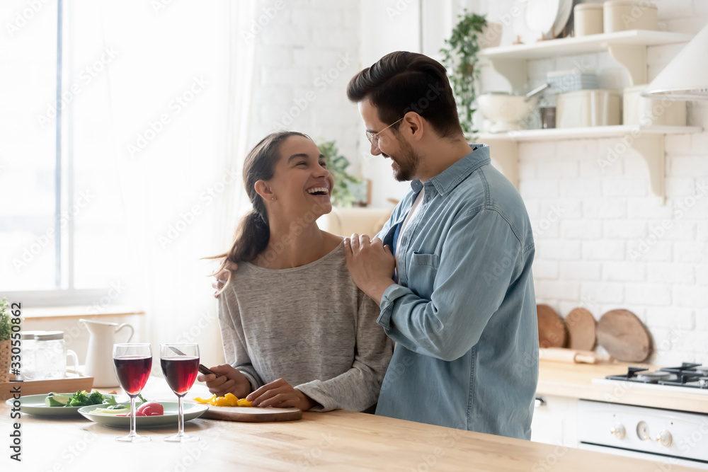 Happy loving couple preparing dinner in modern kitchen together, smiling husband hugging beautiful young wife cutting vegetables for salad, family enjoying free weekend together, having fun Happy loving couple preparing dinner in modern kitchen together, smiling husband hugging beautiful young wife cutting vegetables for salad, family enjoying free weekend together, having fun