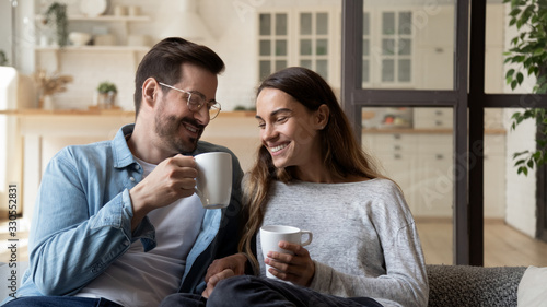 Happy loving couple relaxing on couch, drinking hot beverages, tea or coffee, holding white cups, smiling young man and woman chatting, talking, spending lazy weekend at home, sitting on cozy sofa