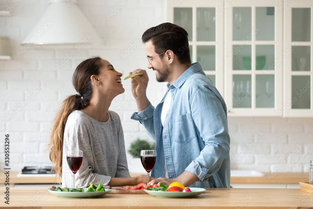 Smiling loving husband feeding wife, young couple enjoying tender moment, romantic date, having fun, eating salad and drinking red wine, happy family spending free weekend in modern kitchen