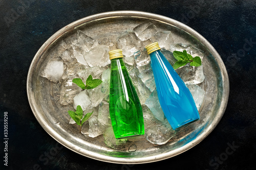 Refreshing bottled drinks, green tarragon lemonade and blue drink on a metal tray with ice cubes and mint leaves. Top view, flat lay.