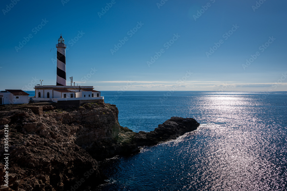 the solid lighthouse on the shore coastline of the island of mallorca, spain, during the late afternonn where sun starts seeting and reflection on the meditereanan sea 