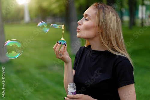 Young beautiful girl inflates soap bubbles in the summer sunny park