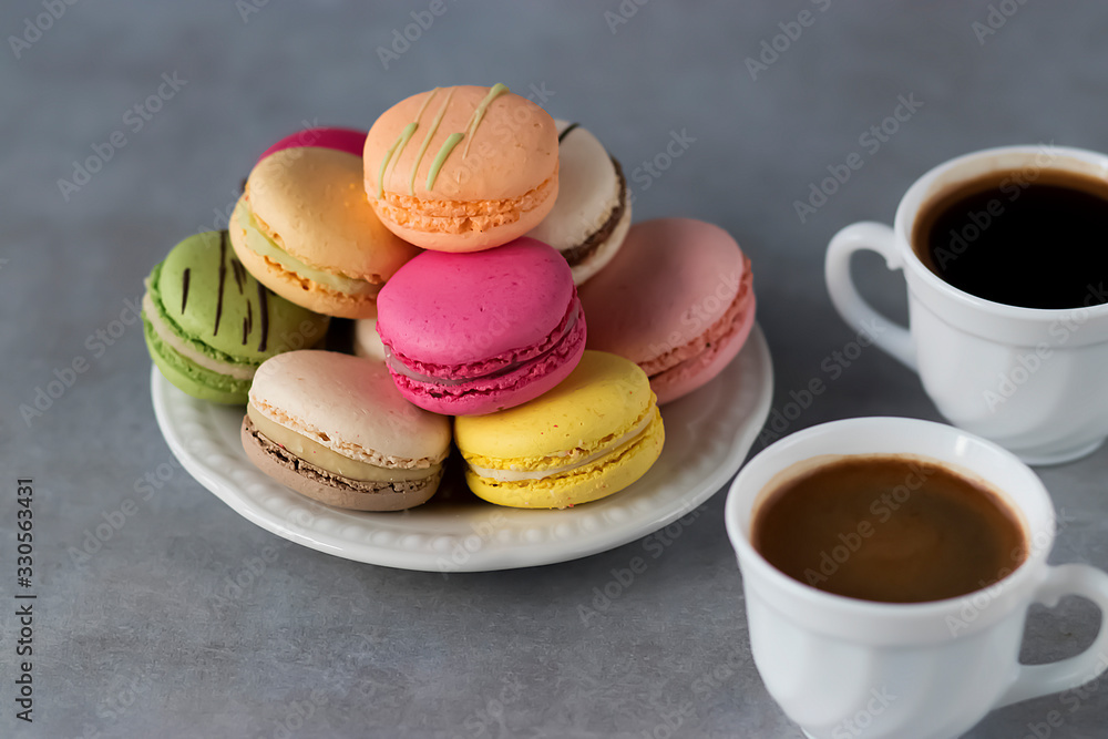 Assorted macaroon cakes in a white plate and two white cups of coffee on a gray background.