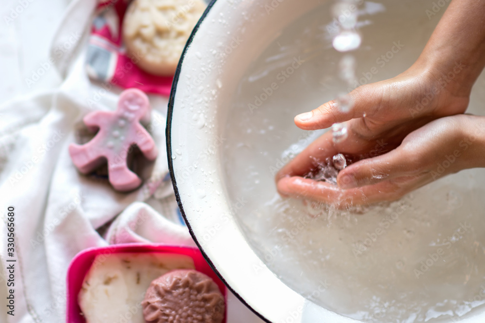Child’s hands under white bowl with water upon water stream, colorful soaps on a white material, cleanliness and hygiene concept Child’s hands under white bowl with water upon water stream, colorful soaps on a white material, cleanliness and hygiene concept