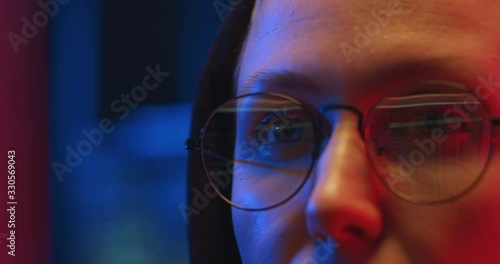 Close up of eyes in glasses of Caucasian woman in hood working at computer screen at night in IT office. Female hacker stealing data in internet or creating and spreading virus. photo