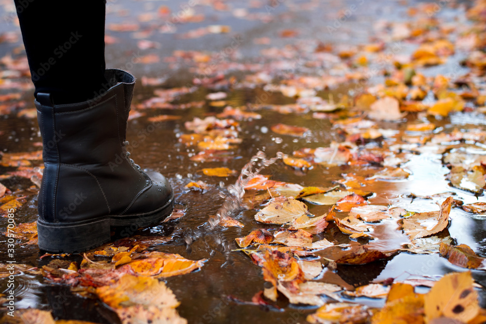 Woman in weather sealed boots going through a puddle with with bright autumn leaves. Concept of autumn rainy weather
