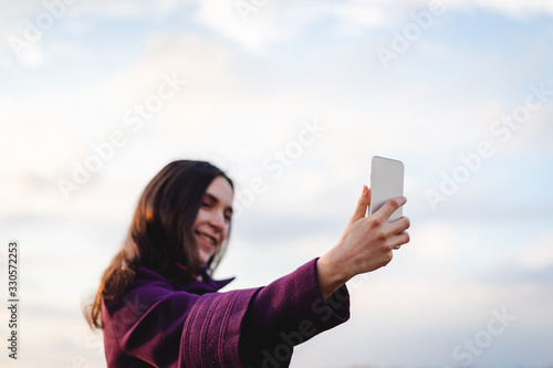 Young caucasian girl with dark hair holding up white mobile phone and taking selfie photo with blue cloudy sky background in susnet light © Khrystyna Pochynok