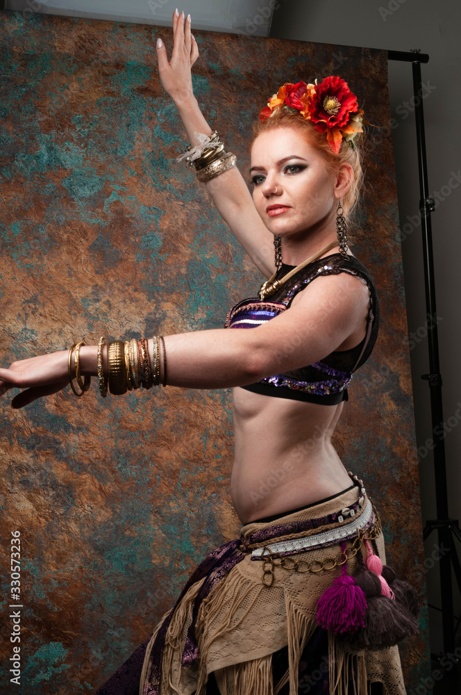 side view of attractive woman in tradition tribal costume with vest, skirt and red floral circlet of flowers stay in pose and looking away in front of teal and orange background in studio side view of attractive woman in tradition tribal costume with vest, skirt and red floral circlet of flowers stay in pose and looking away in front of teal and orange background in studio