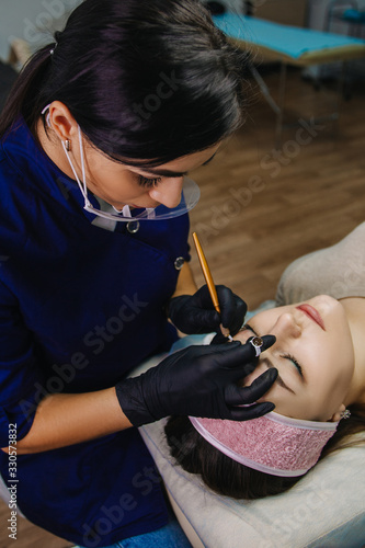 The girl’s face is a profile with her eyes closed, in the background a microblading master in blue uniform and black gloves draws paint from a ring on a needle.