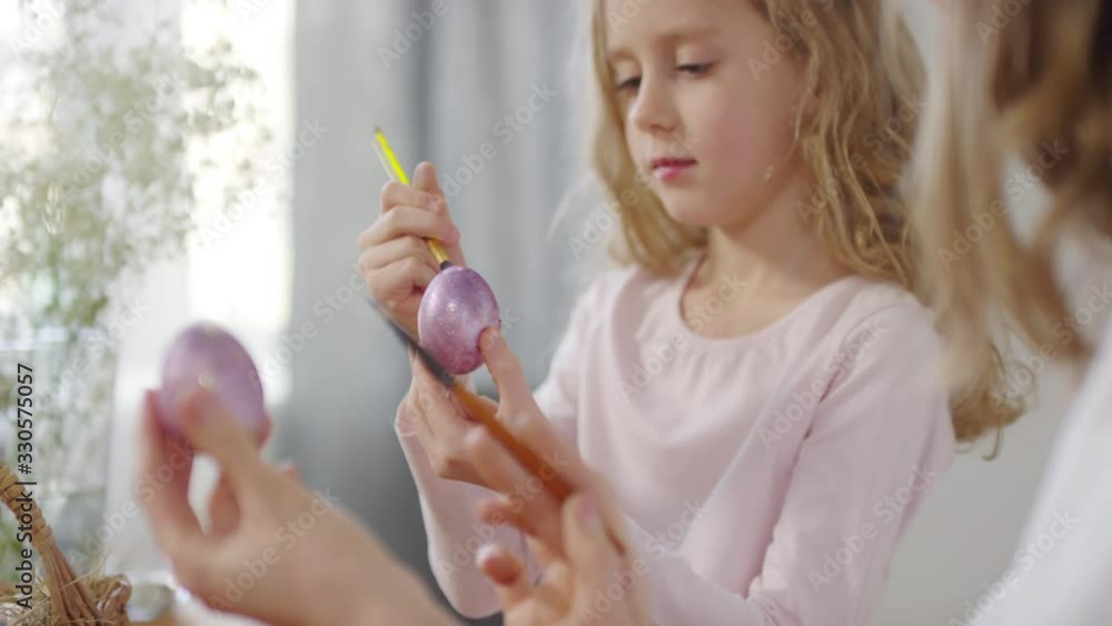 Handheld shot of happy young woman and cute girl talking and putting glitter on purple Easter eggs Handheld shot of happy young woman and cute girl talking and putting glitter on purple Easter eggs