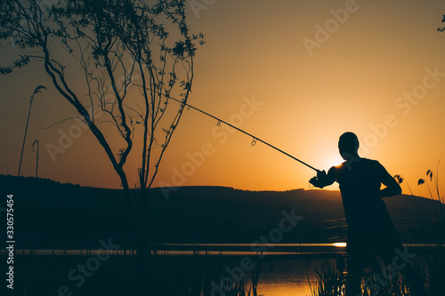 Silhouette of a fisherman fishing on a lake at sunset © belyaaa