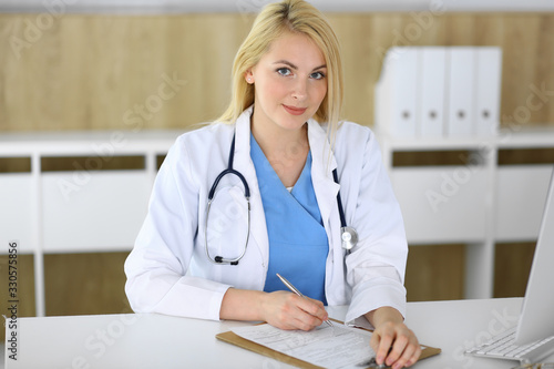 Doctor woman at work while sitting at the desk in hospital or clinic. Blonde cheerful physician filling up medical records form. Data and best service in medicine and healthcare