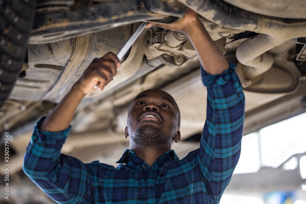 African male car mechanic standing under the car on the lift in the service center, repairs the chassis by tightening the nuts African male car mechanic standing under the car on the lift in the service center, repairs the chassis by tightening the nuts