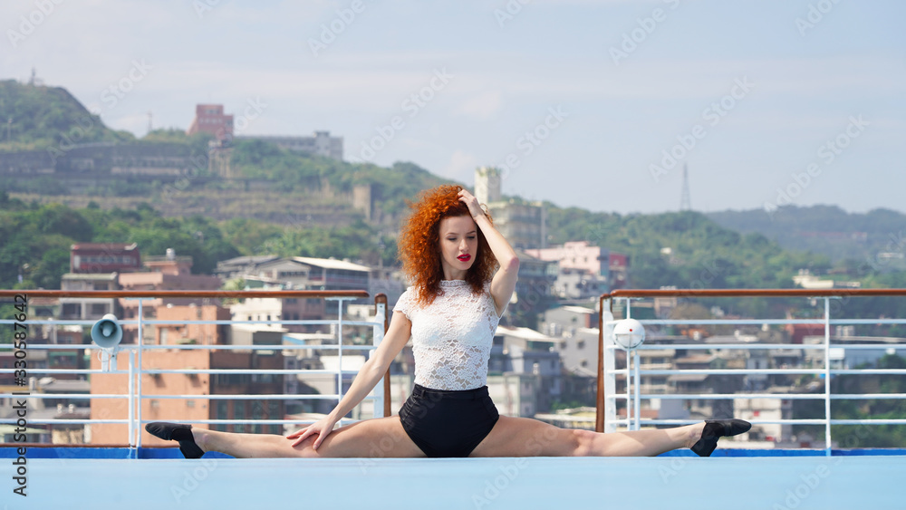 Slim beautiful girl dancer on the observation deck does gymnastic twine. red-haired curly-haired European girl on a blue podium sits in a transverse twine in sun rays on background of city. emotions