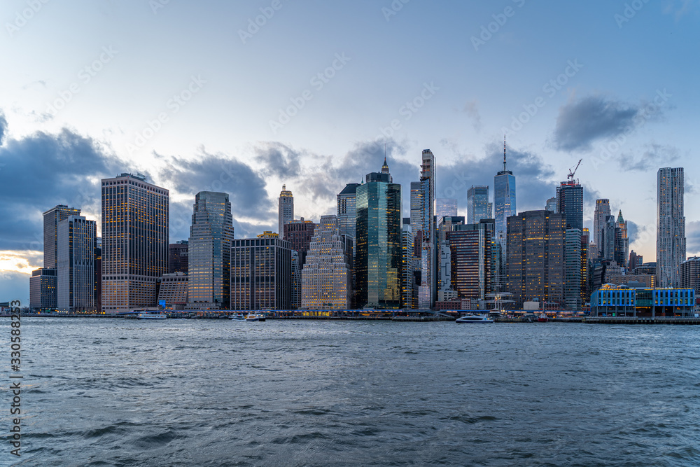 Fototapeta premium New York City Manhattan Skyline at dusk with lights on and clouds from Brooklyn bridge park over the river Hudson after Sunset. Freedom Tower Financial District one world trade center night life 