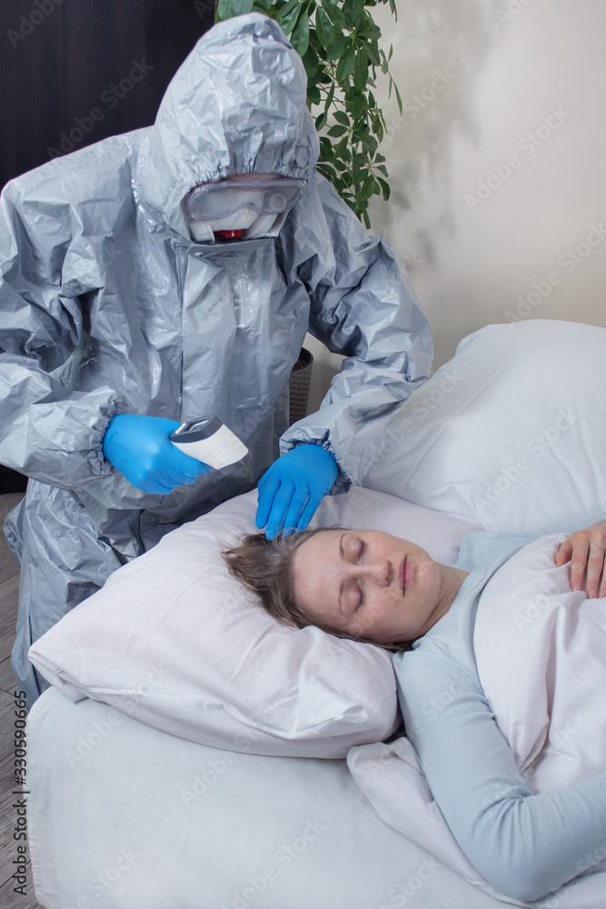 Osoba ubrana w kombinezon ochronny, maskę i gogle ochronne mierzy temperaturę chorej kobiecie leżącej w swoim łóżku. Walka z epidemią.